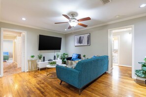 Una sala de estar con un sofá color turquesa, suelo de madera, un ventilador de techo, un televisor montado y una puerta que da a otra habitación.