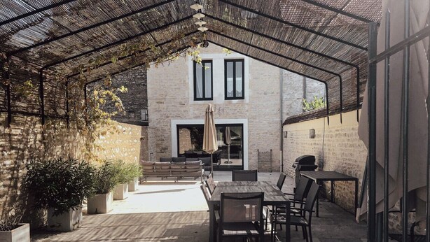 A covered outdoor patio with a dining area, stone walls, and a view of a building with two windows.
