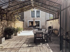 Une terrasse extérieure couverte, dotée d’un espace repas, de murs en pierre et offrant une vue sur un bâtiment avec deux fenêtres.