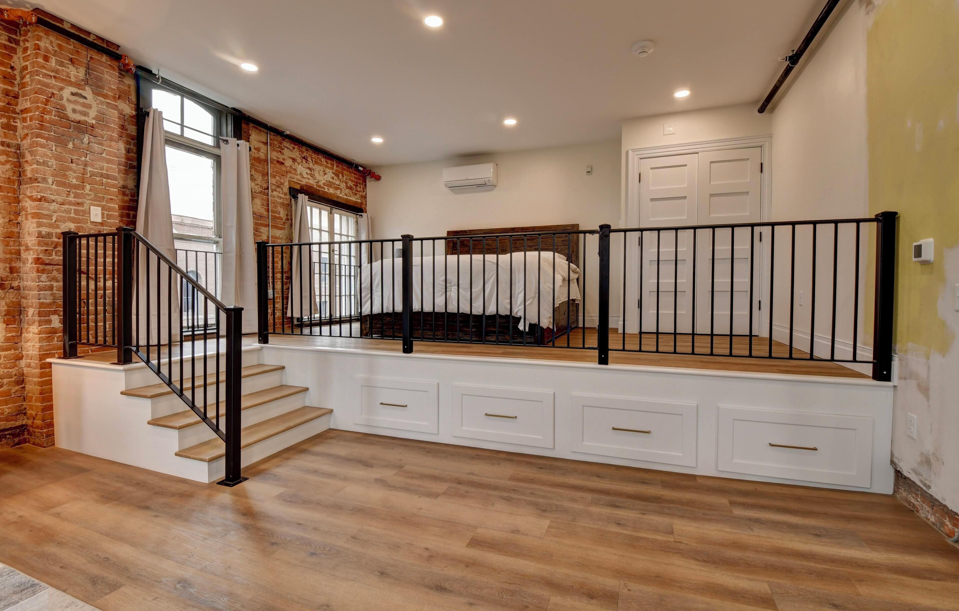 Une chambre avec un lit, un escalier doté de rampes noires et un mur blanc avec une fenêtre.