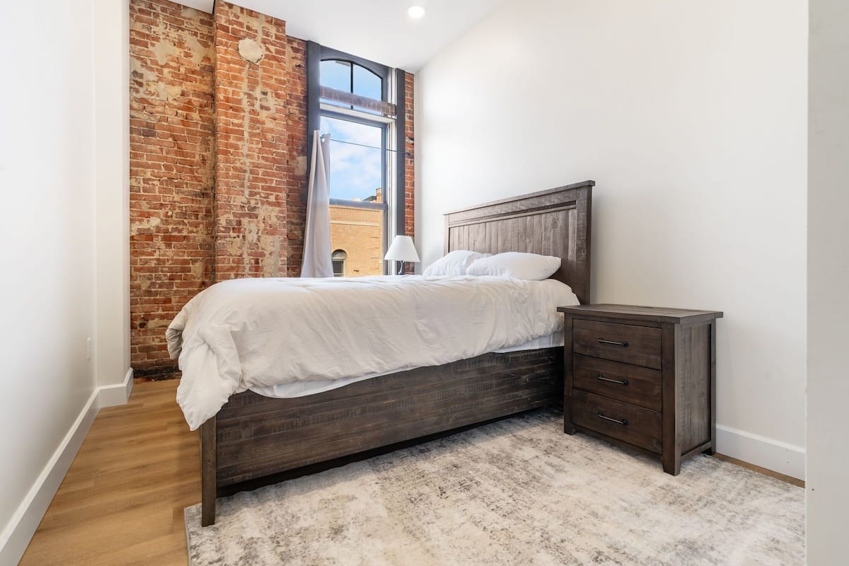 A bedroom with a brick wall, a large window, a bed with white bedding, a bedside table, and a wooden floor.