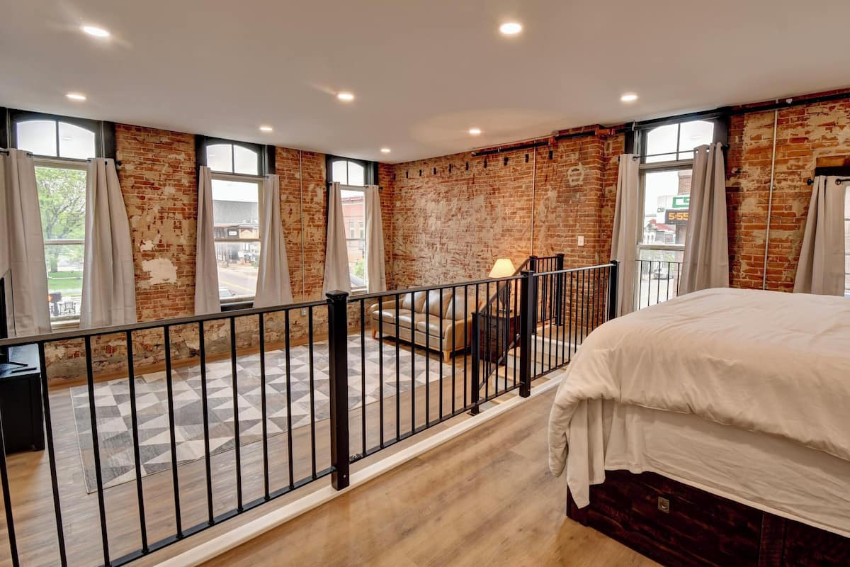 A bedroom with a large bed, a view of the street through a window, a brick wall, and a balcony with a railing.