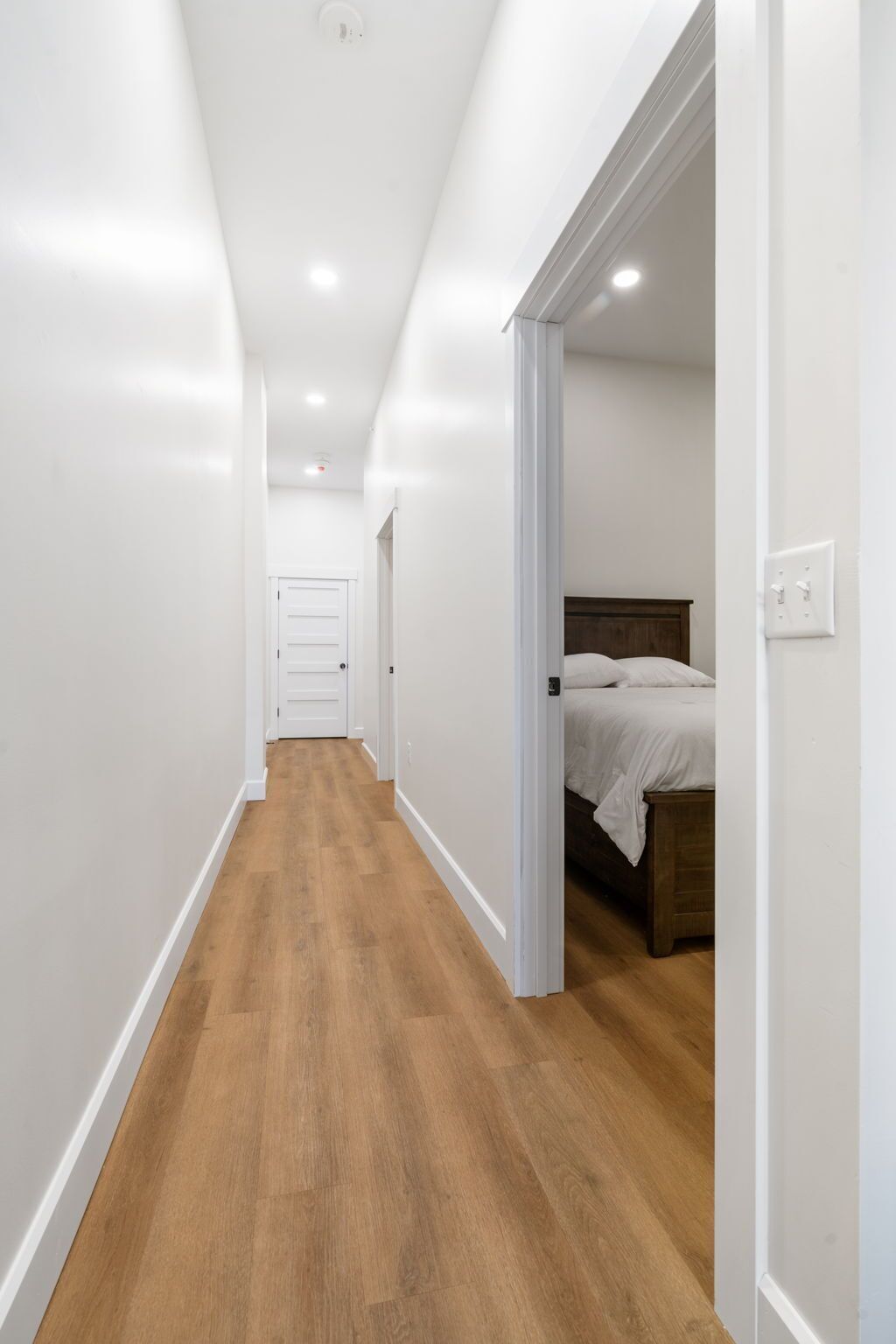 A narrow hallway with wooden flooring leading to a bedroom with a bed and a nightstand.