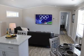 Un salon moderne doté d’un téléviseur à écran plat affichant les anneaux olympiques, d’un canapé noir, d’un coin repas avec une table dressée pour deux personnes et d’un espace cuisine avec un panier de fruits posé sur le comptoir.