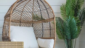 Una silla colgante de mimbre con cojines blancos, una mesa de vidrio y una planta en maceta.