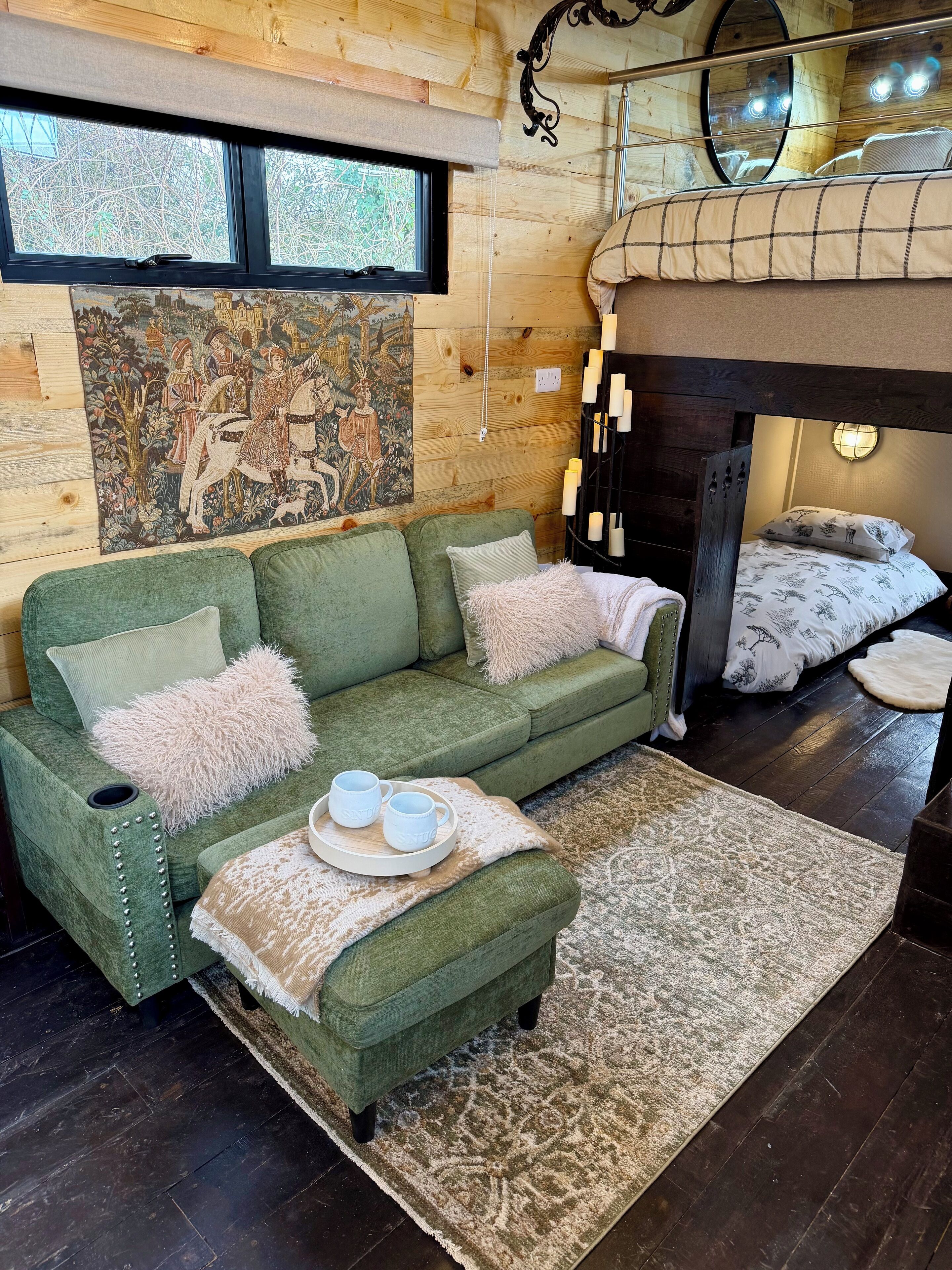 Una habitación acogedora con un sofá verde, una mesita con dos tazas, un tapiz decorativo y una cama litera con un espejo.