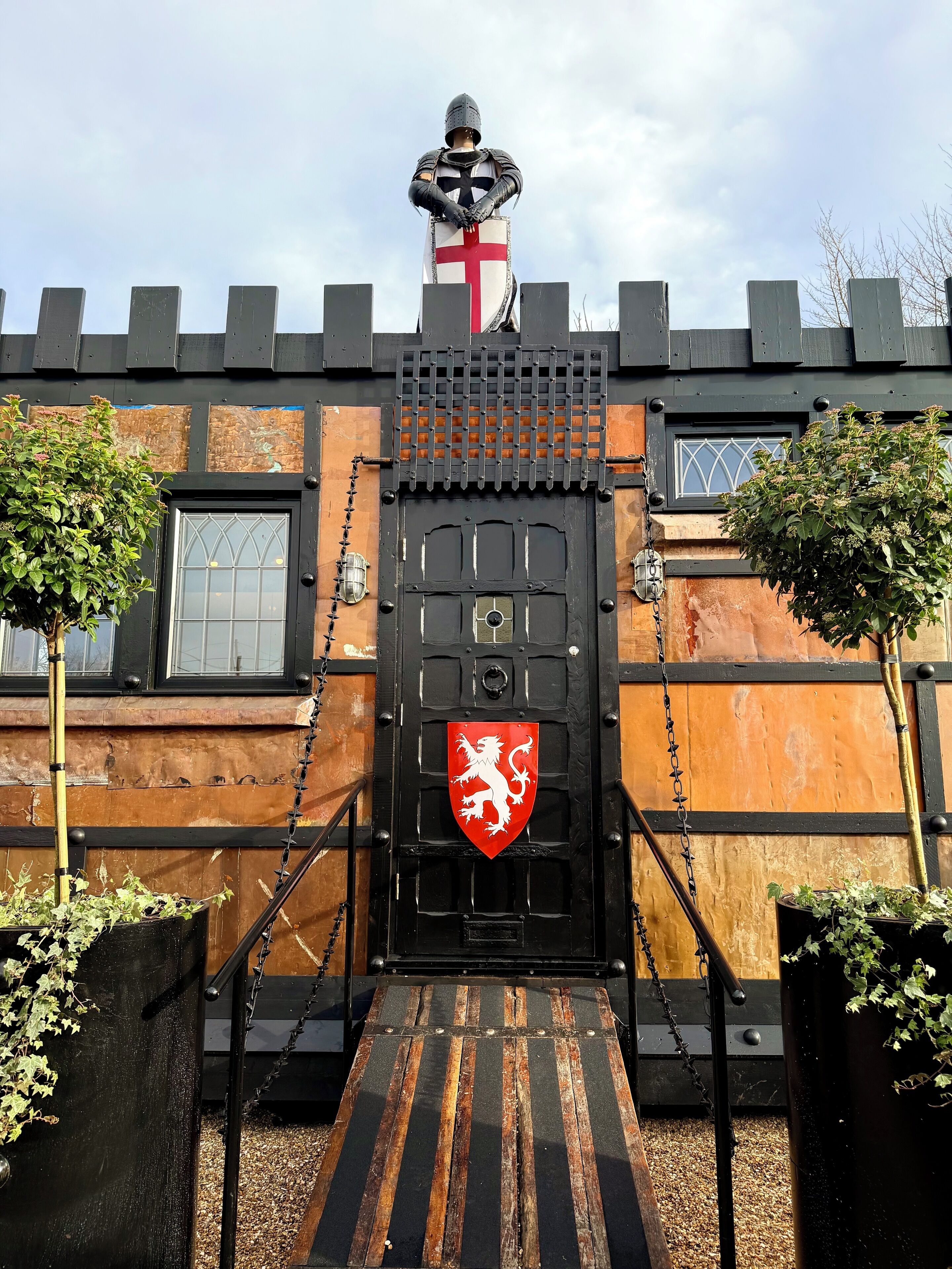 A medieval-themed building with a black gate, a knight statue, and a red shield emblem.