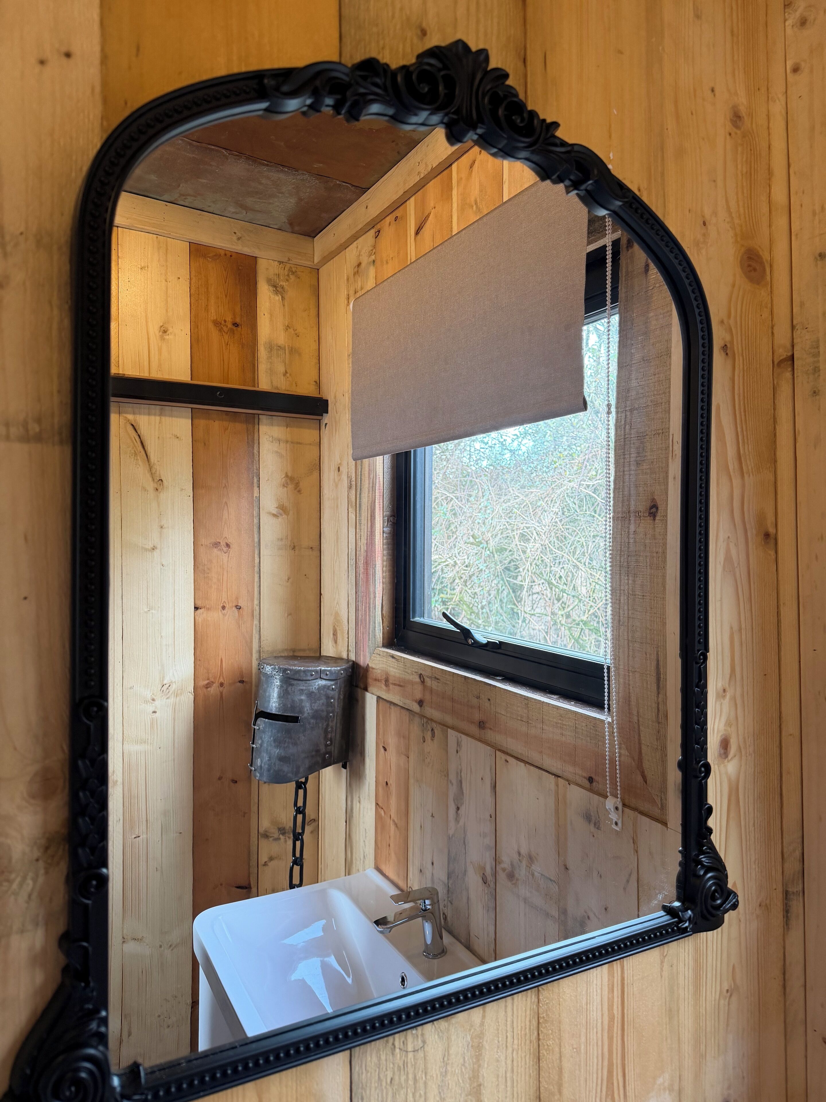 Interieur van een houten hutje met een badkamer, inclusief een wit wastafeltje, een raam met een zonwering en een spiegel met een sierlijk frame.