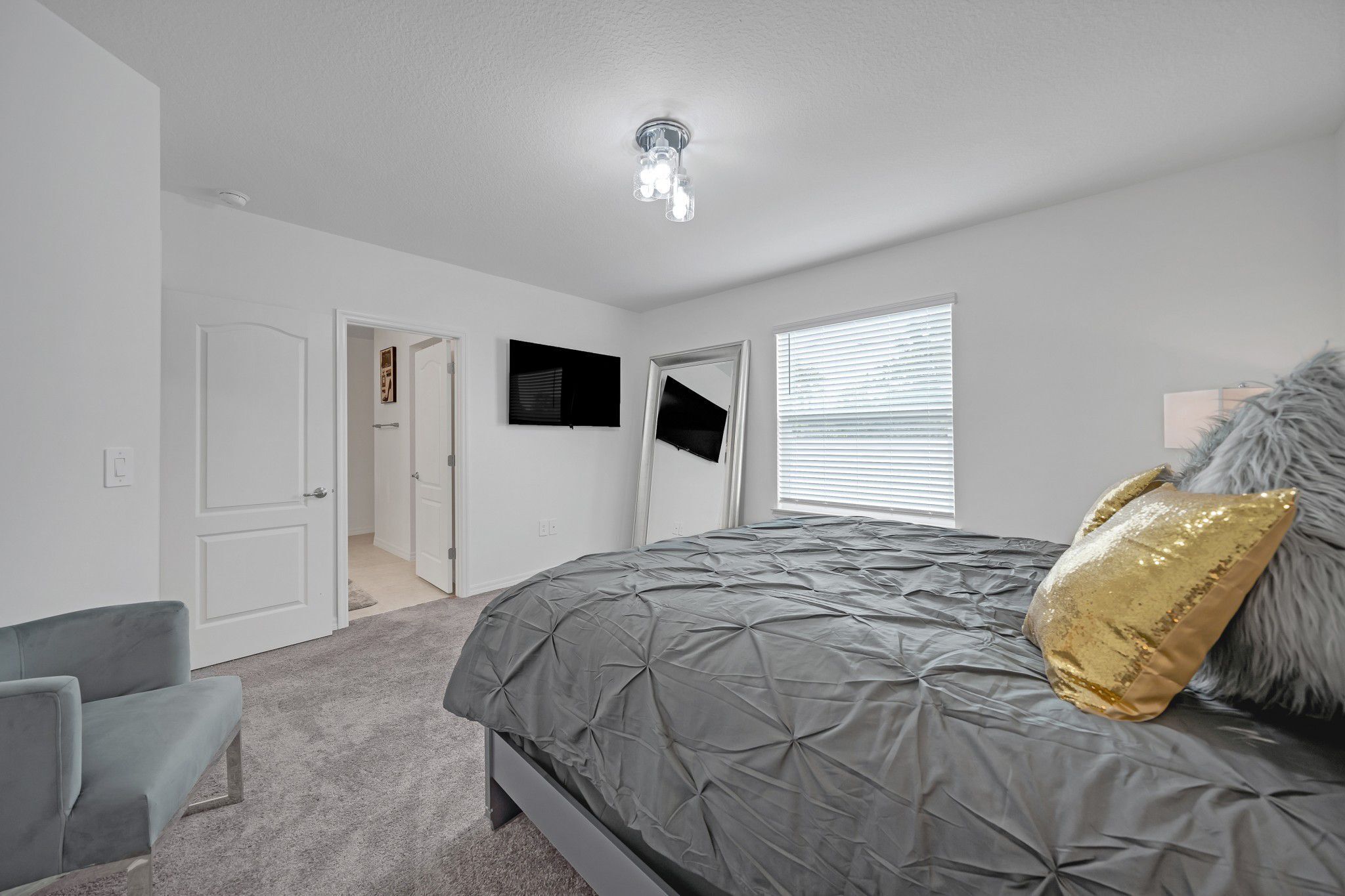 Quarto com cama, sofá cinzento, porta e luminária no teto.