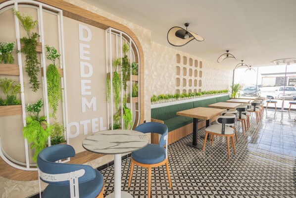 Un hall de hotel con diseño moderno, que presenta una zona de estar con mesas y sillas, una pared decorativa con plantas y una señalización visible que indica "DEDEM HOTEL".