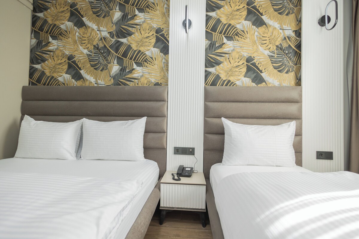 Habitación de hotel con dos camas, una mesita de noche con teléfono y un papel tapiz con motivos de hojas tropicales.
