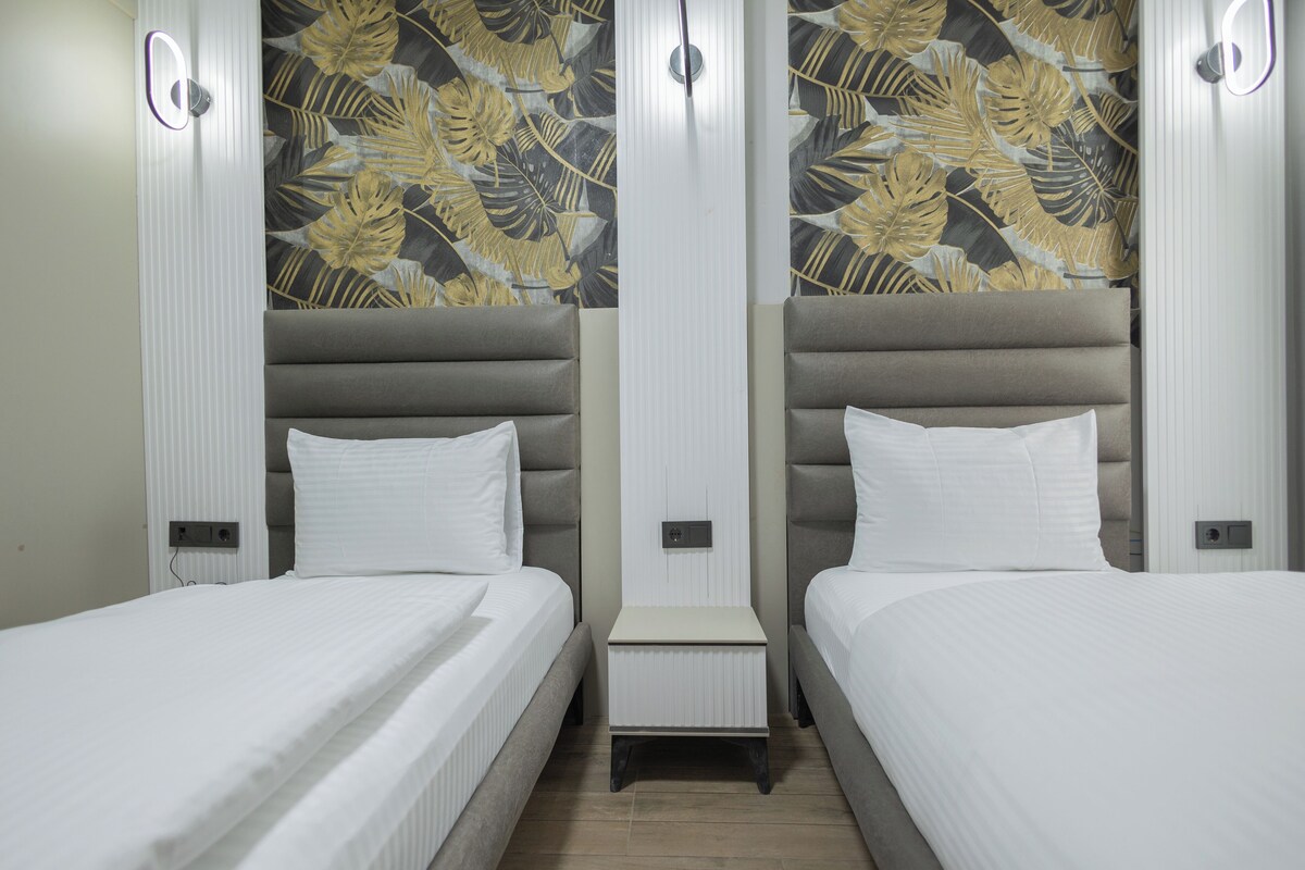 Habitación de hotel con dos camas, una mesita de noche pequeña y una pared con un diseño de hojas.