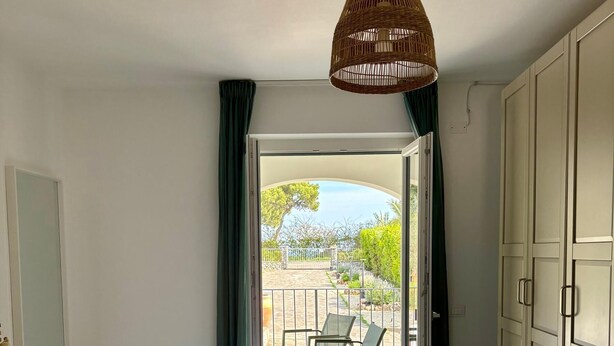 Quarto com cama, luminária pendente e vista através de uma porta aberta para área externa com cadeiras e vegetação.