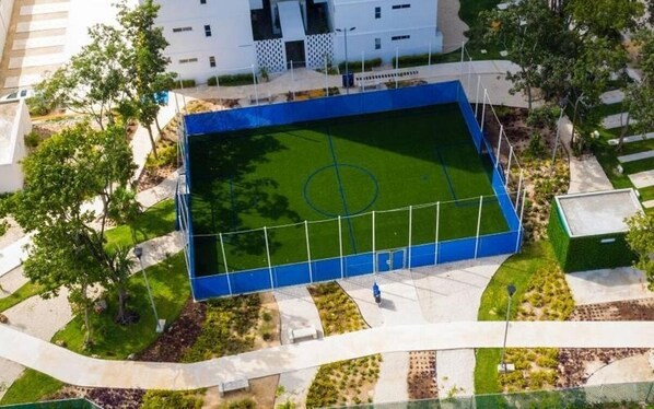 An aerial view of a sports field with a blue fence, surrounded by landscaped areas and a building in the background. - Elegant 2BR/2BA Retreat | Pools | 24/7 Sec (Cancún)