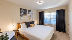 Une chambre à coucher avec un grand lit, une table de chevet, une lampe et une fenêtre avec des rideaux.