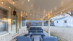Une terrasse extérieure dotée d’un espace salon couvert, de guirlandes lumineuses et offrant une vue sur une maison en arrière-plan.