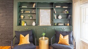 Une pièce comprenant deux fauteuils, une table ronde et une bibliothèque verte remplie de livres et d’objets décoratifs.