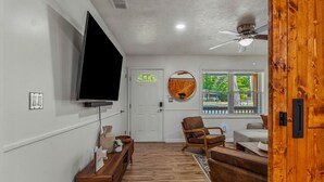 A living room with a flat-screen TV mounted on the wall, a wooden floor, a ceiling fan, and a large window with a view of greenery outside. - Butterfly Bungalow is a 4BR/2BA home with outdoor terrace and grill (Columbus)