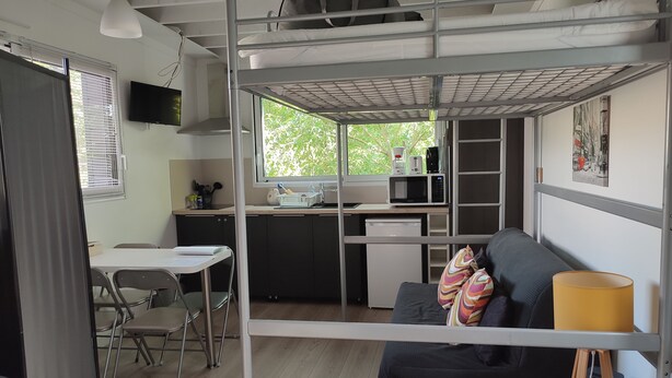 A compact living space with a bunk bed, kitchenette, dining area, and a window with a view of greenery. - Fully-equipped 1 room furnished studio without partitions (Chavagnes-en-Paillers)