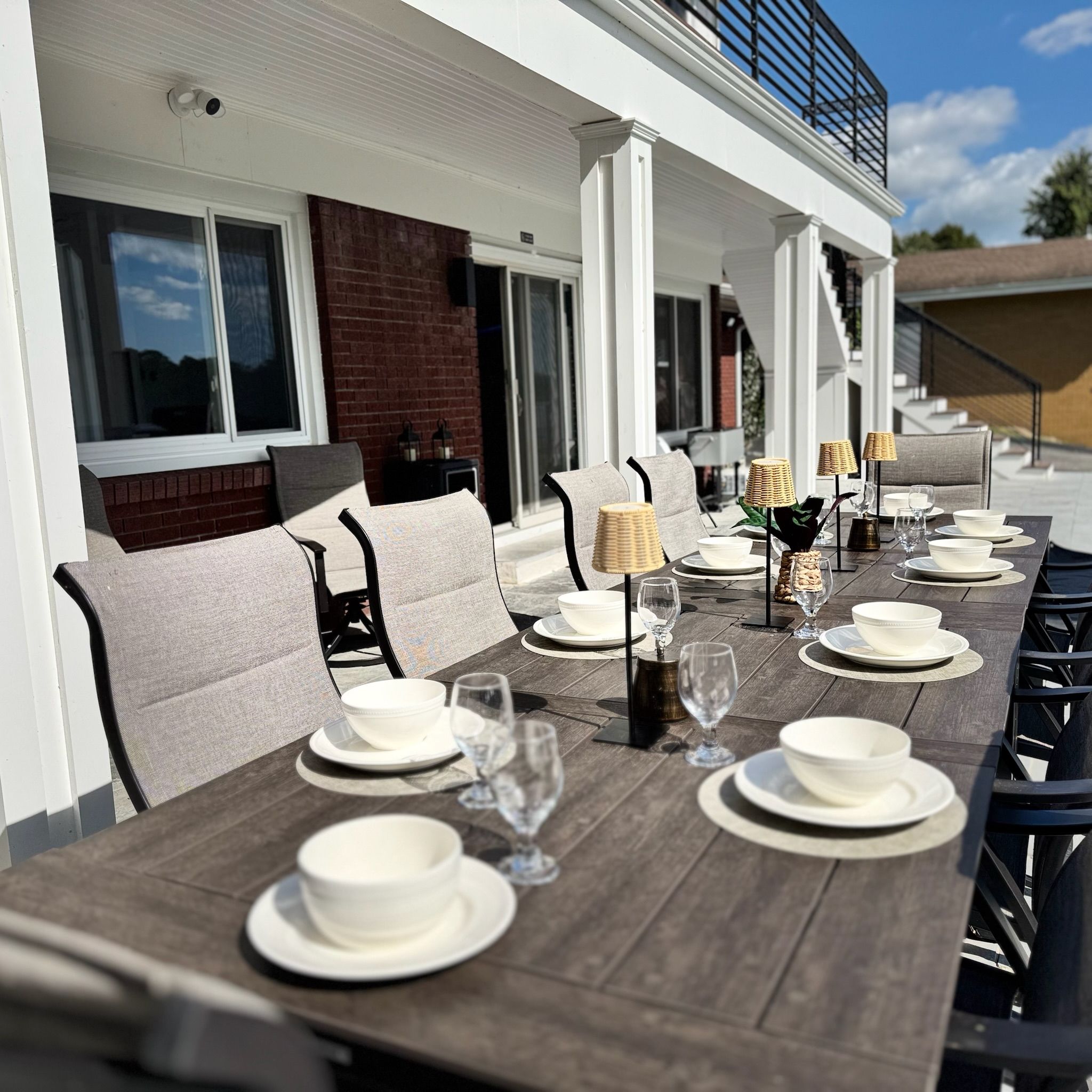 An outdoor dining area with a long wooden table set for a meal, surrounded by cushioned chairs.