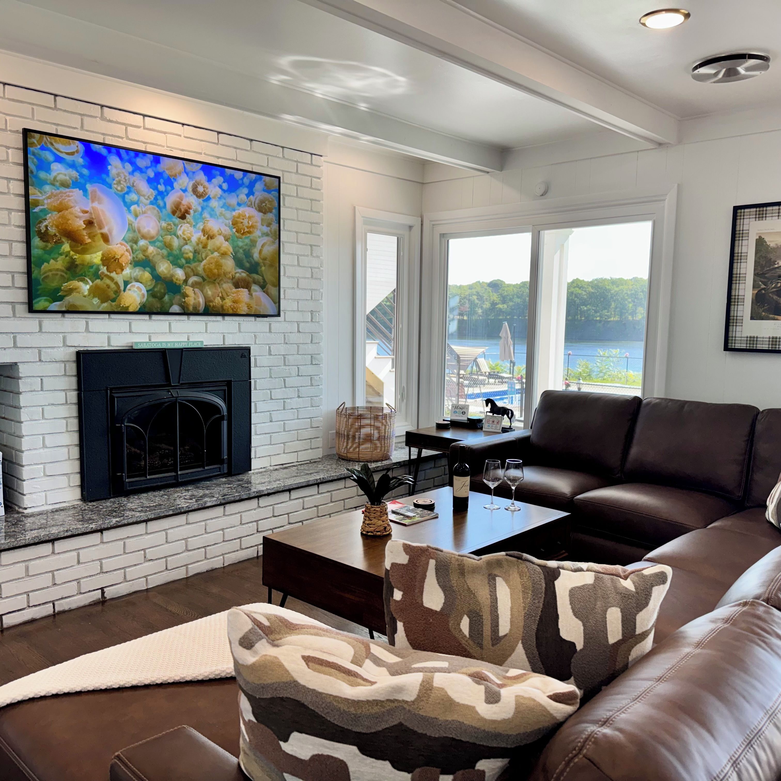 A living room with a fireplace, a large TV mounted on a brick wall, a brown leather couch, and a view of a lake through large windows.