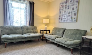 Une pièce avec deux canapés verts, une petite table avec une lampe, une fenêtre ornée de rideaux à motifs et un tableau abstrait accroché au mur.