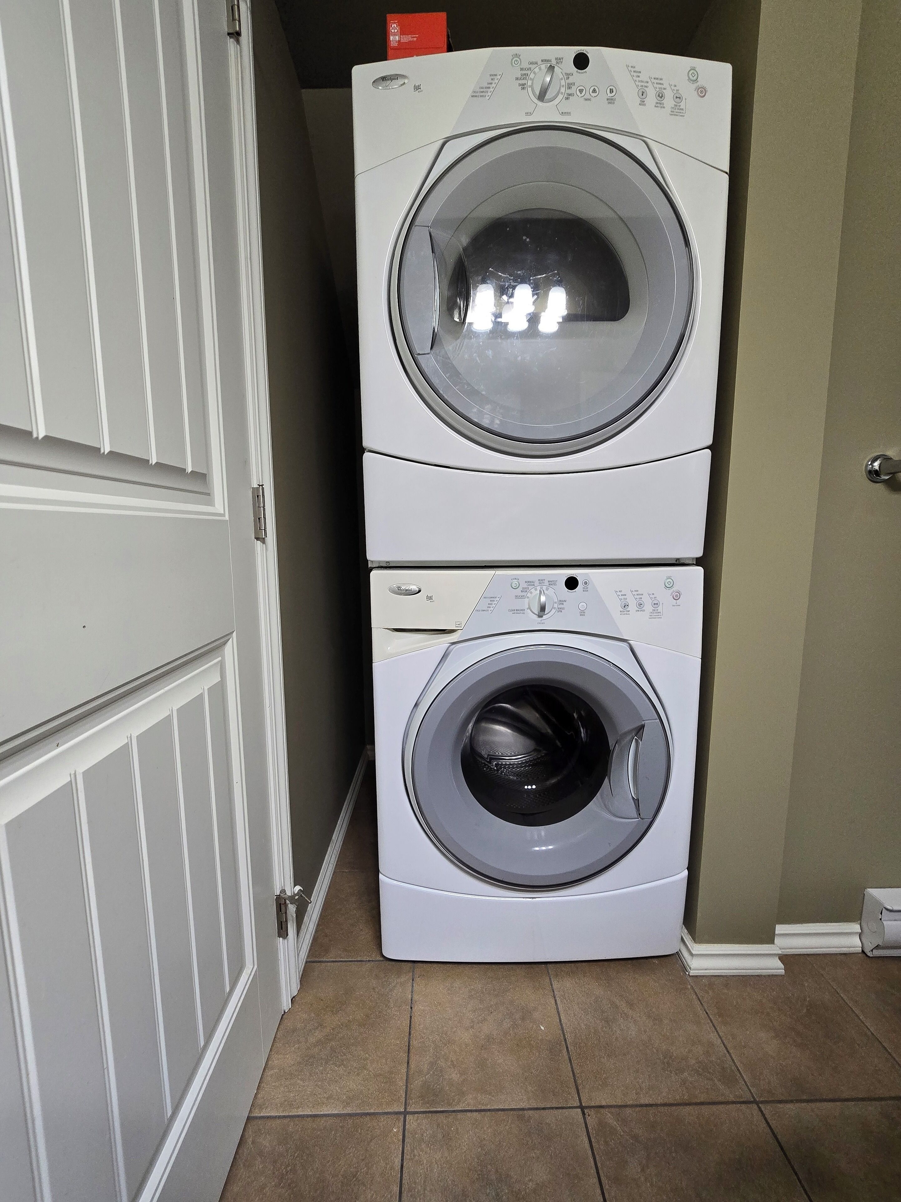 Une machine à laver et un sèche-linge superposés, installés dans un couloir avec une porte fermée et un sol carrelé.