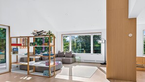 A modern living room with a sectional sofa, a wooden bookshelf, and large windows offering a view of greenery. - Dartmouth Graduation—Luxury MCM Home in Hanover (Hanover)