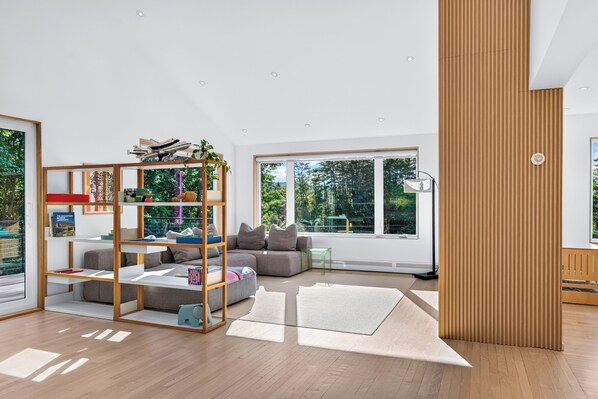 A modern living room with a sectional sofa, a wooden bookshelf, and large windows offering a view of greenery. - Dartmouth Graduation—Luxury MCM Home in Hanover (Hanover)