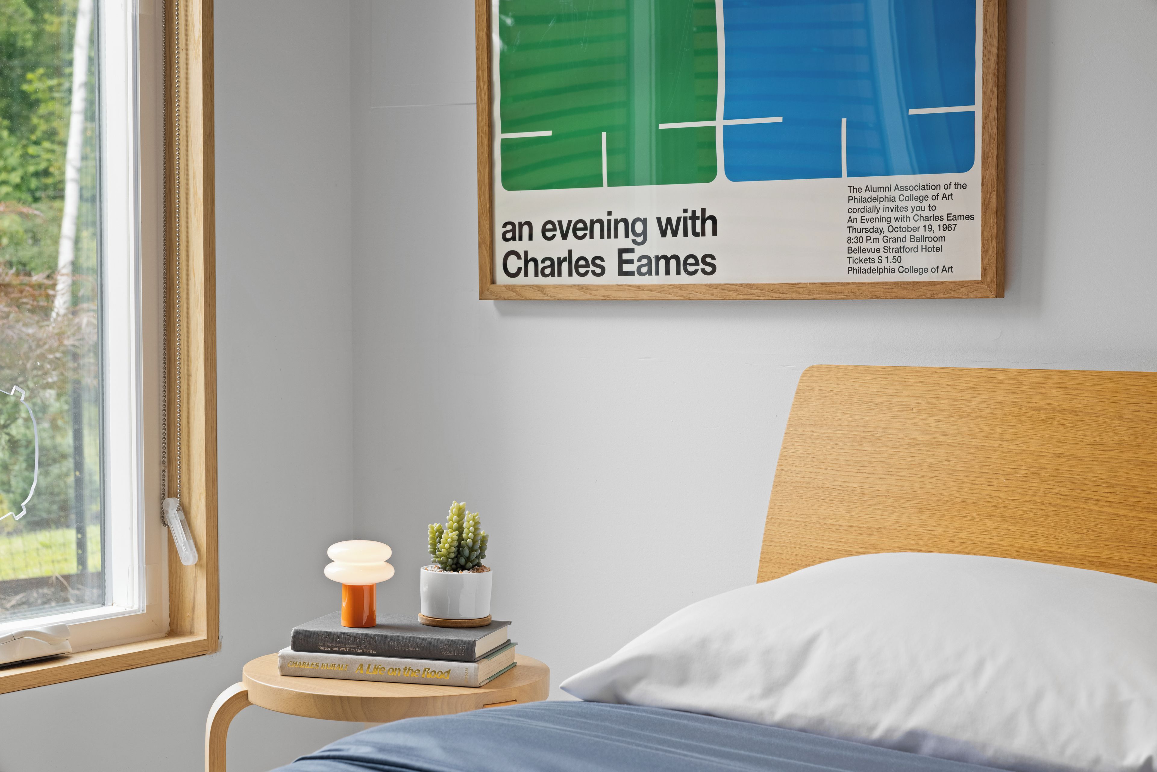 Een keurig opgemaakt slaapkamer met een bed, een nachtkastje met boeken en een kleine plant, een lijst met een poster aan de muur en een raam met uitzicht op groen.
