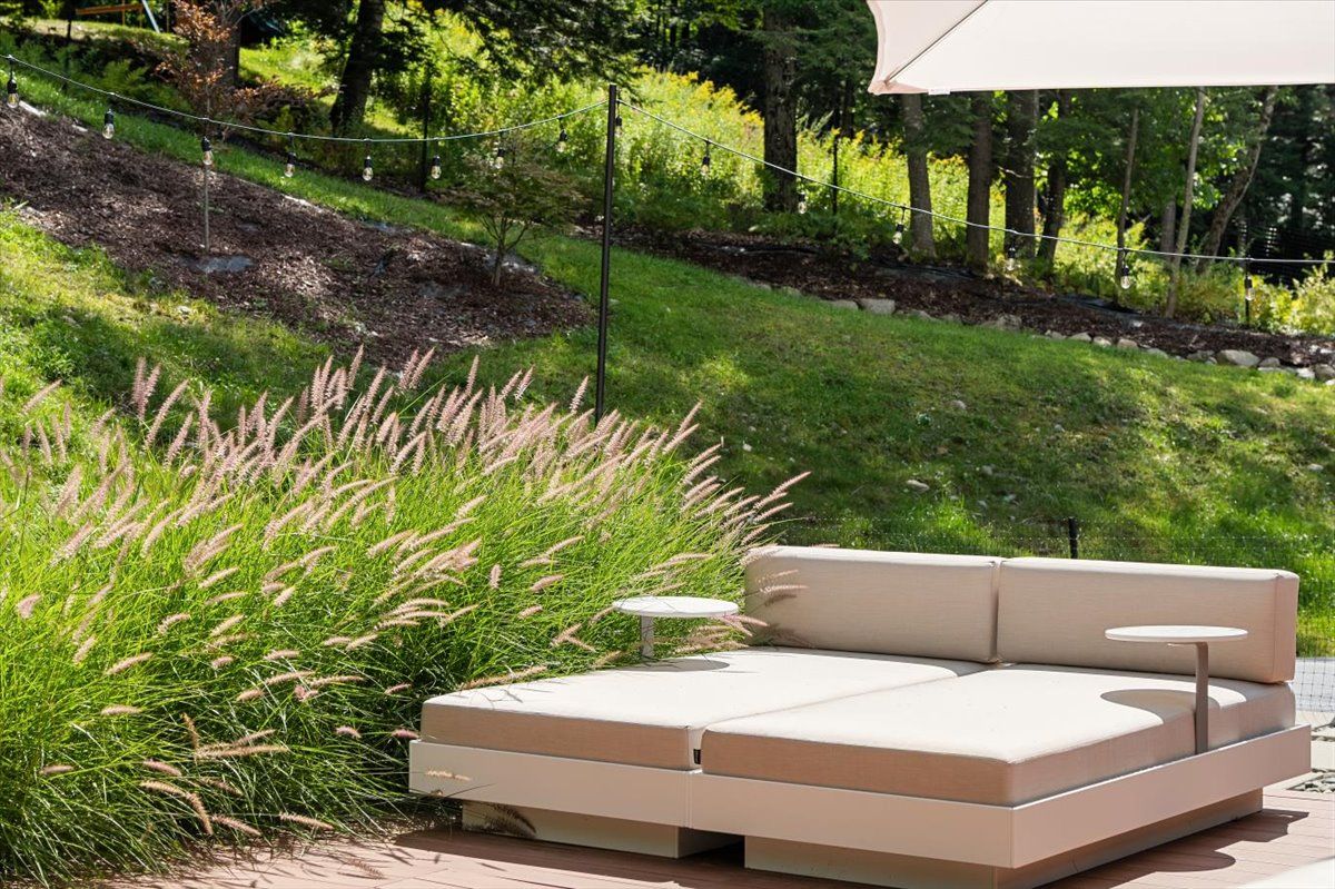 An outdoor seating area with a modern sofa and two small tables, surrounded by lush greenery and a string of lights.