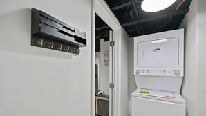 Uma lavanderia com máquina de lavar e secadora brancas, aquecedor fixado na parede e porta que dá para outro cômodo.