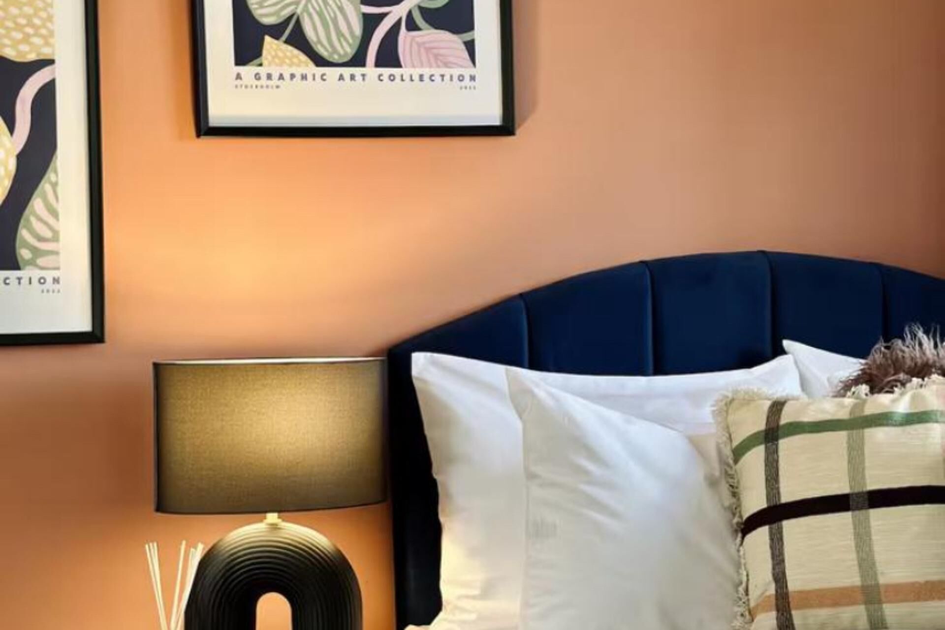 A bedroom with a dark blue headboard, a bedside lamp, and a framed artwork on the wall.