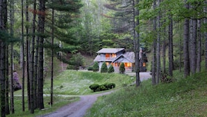 Una casa con tetto spiovente, circondata da alberi alti e un vialetto in ghiaia che vi conduce.