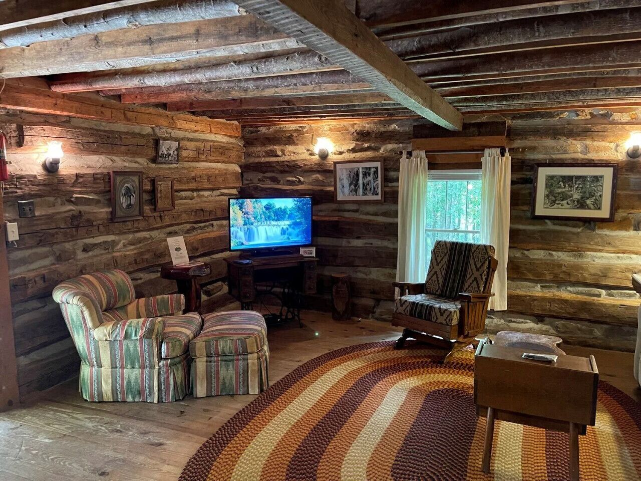 A rustic cabin interior with wooden walls, a television, striped armchairs, a patterned rug, and a wooden coffee table.