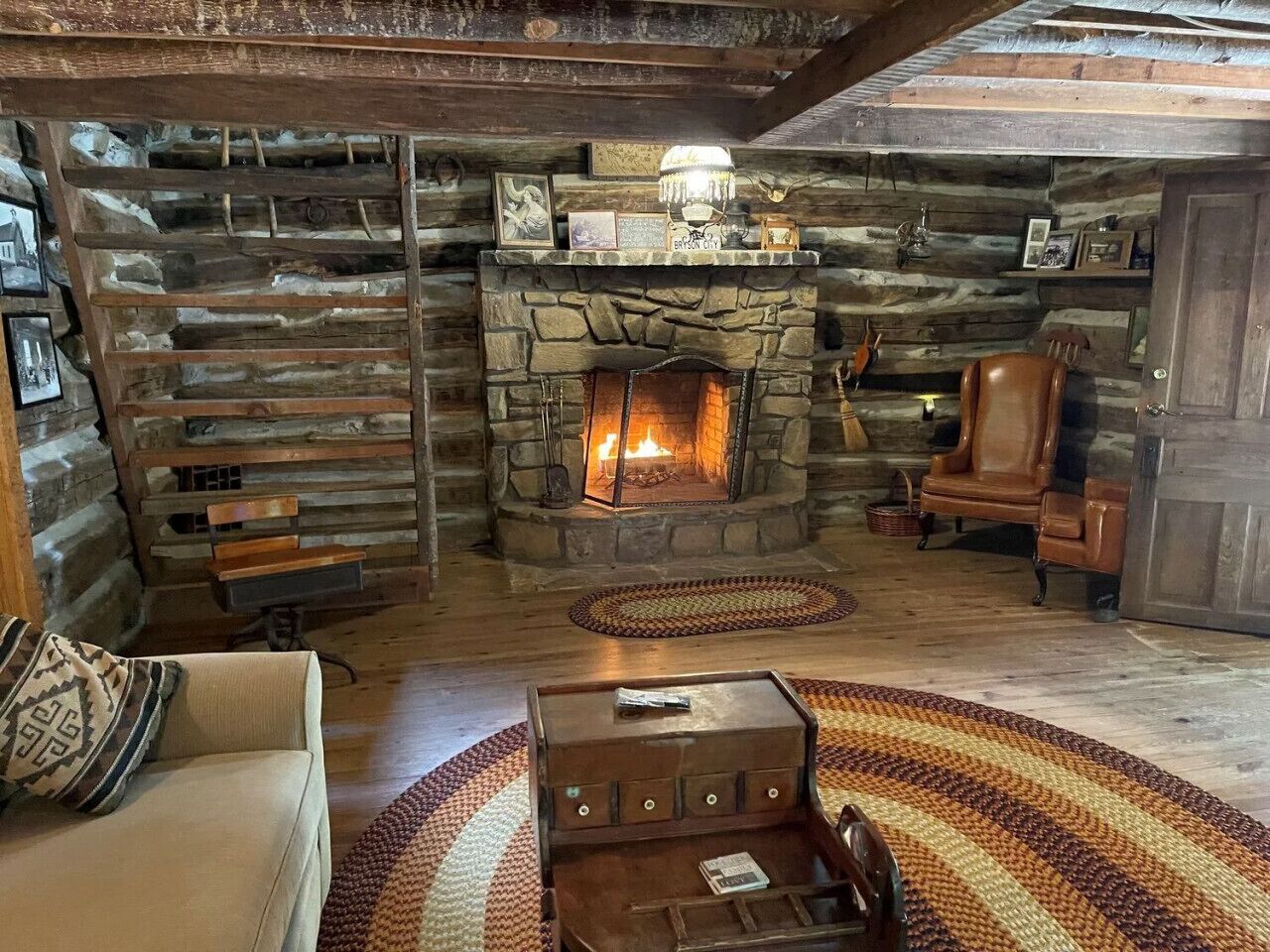 A cozy log cabin interior with a stone fireplace, wooden benches, a leather armchair, and a patterned rug.
