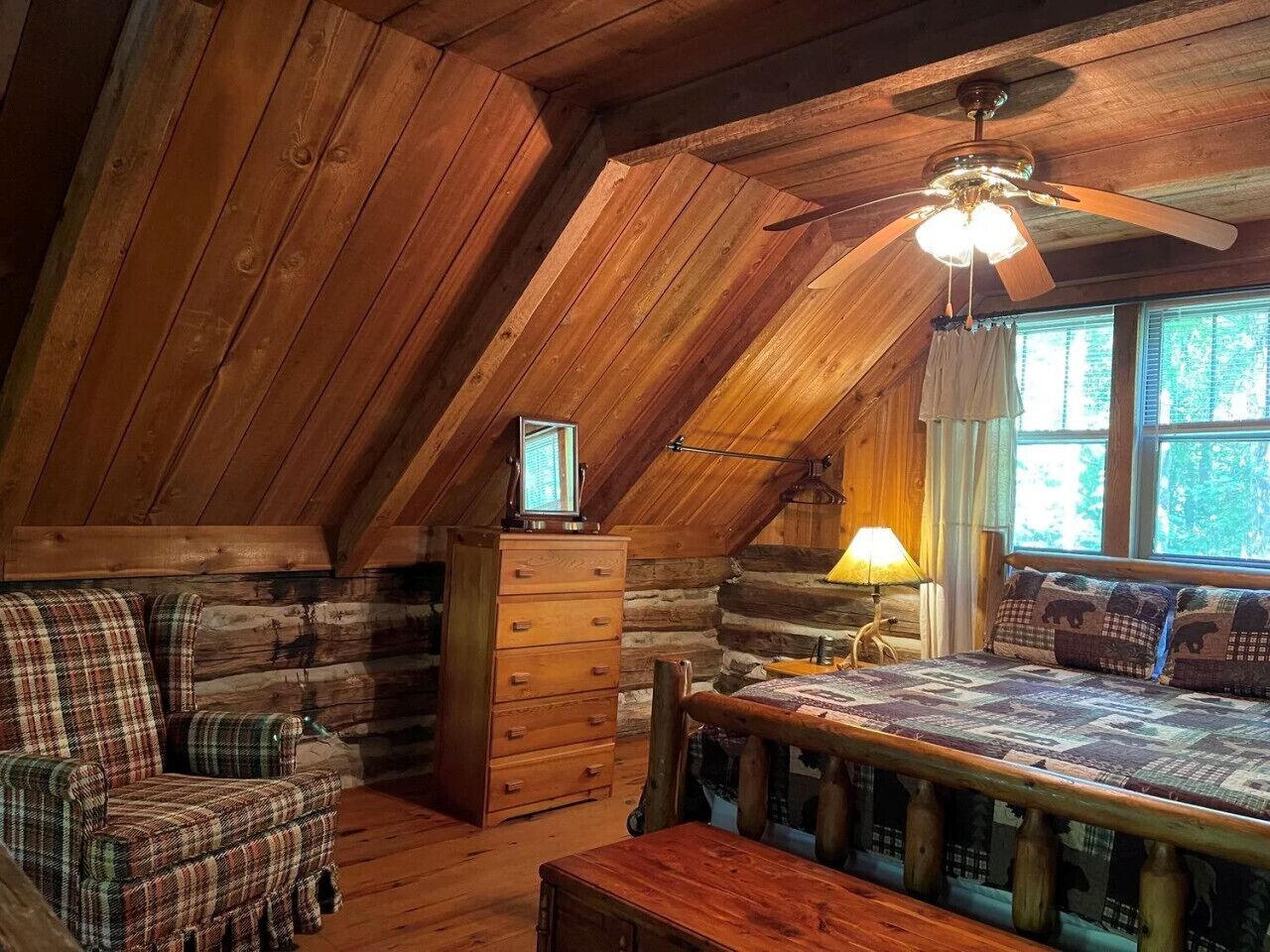 A cozy log cabin bedroom with a wooden ceiling, a bed with a quilt, a dresser, a chair, and a window with curtains.