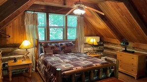 Un dormitorio acogedor en el ático con techo de madera, una cama con colcha estampada, dos mesitas de noche con lámparas, un aparador de madera y una ventana con cortinas.