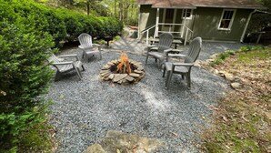 A cozy outdoor seating area with a fire pit surrounded by Adirondack chairs, set against a backdrop of a rustic house and lush greenery. - Private fire pit, creek: Bryson city (Bryson City)