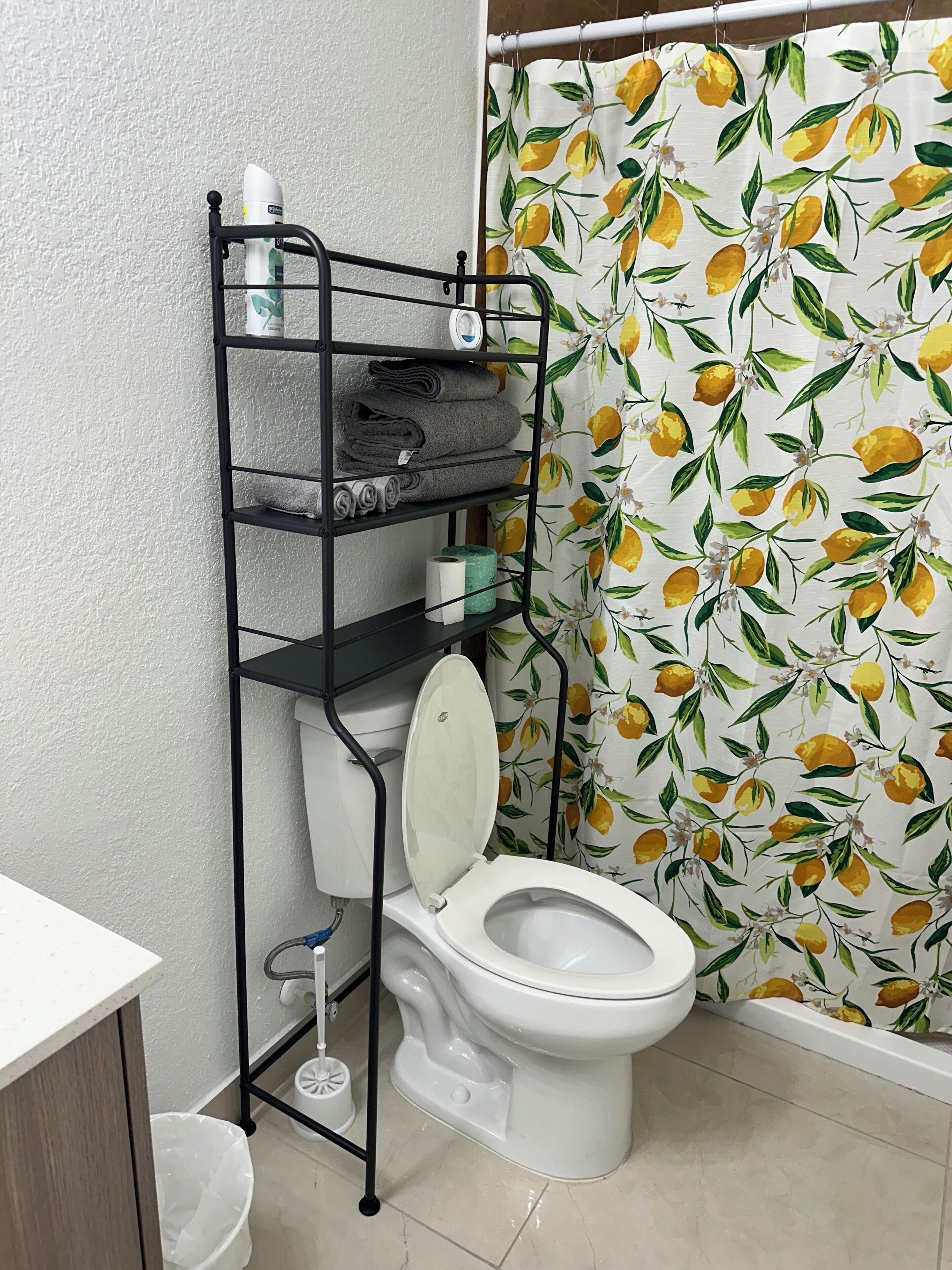 A bathroom with a toilet, a shelf unit with towels, and a shower curtain with a lemon pattern.