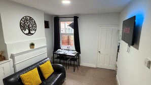 A compact living space with a black leather sofa, a dining table with chairs, a TV mounted on the wall, and a door leading to another room. - Amazing Garden Apartment - Miami Lodge (Reading)