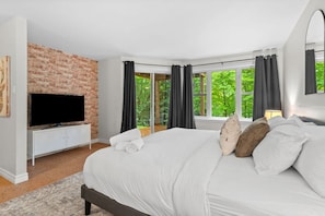A modern bedroom with a brick accent wall, a large bed, a television, and a view of greenery through the windows. - Boisé 4-Story Ski-In/Ski-Out w/ Sauna (Mont-Tremblant)