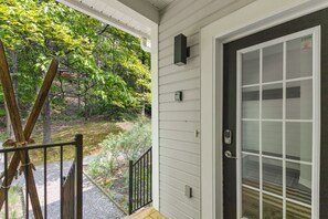 Varanda com vista para uma área arborizada, porta com olho mágico e pequena luminária externa.