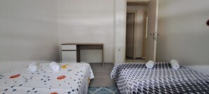 Ein kleines Hotelzimmer mit zwei Einzelbetten, einem Schreibtisch und einer Tür zu einem weiteren Raum.