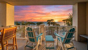 Un balcon avec des chaises et une table en bois, donnant sur une plage bordée de palmiers et illuminée par le coucher de soleil.