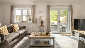 Un salon avec un canapé, une table basse et un téléviseur. Il y a des fenêtres avec des rideaux, une lampe et une vue sur la verdure à l’extérieur.