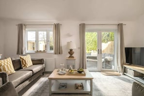A living room with a sofa, coffee table, and a TV. There are windows with curtains, a lamp, and a view of greenery outside. - New Market - Modern Stay in Cotswolds with Hot Tub (Gloucestershire)