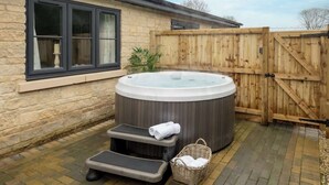 A hot tub in a backyard with a stone wall, wooden fence, and a window with curtains. - Goodwood - Modern Stay in Cotswolds with Hot Tub (South Cerney)