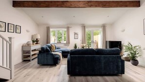 A spacious living room with a large dark blue sofa, a smaller sofa, and a television. There are windows with curtains, a wooden ceiling beam, and a staircase on the left. - Goodwood - Modern Stay in Cotswolds with Hot Tub (South Cerney)