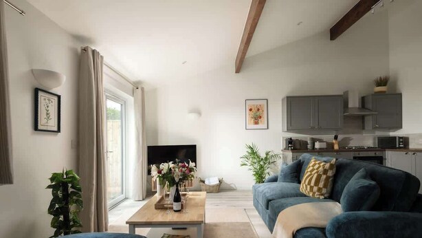 A modern living room with a sofa, coffee table, and TV. There is a kitchen area with cabinets and a plant. - Goodwood - Modern Stay in Cotswolds with Hot Tub (South Cerney)